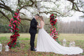 Couple sharing a kiss under a floral arch with red and pink flowers, surrounded by trees and lanterns.
