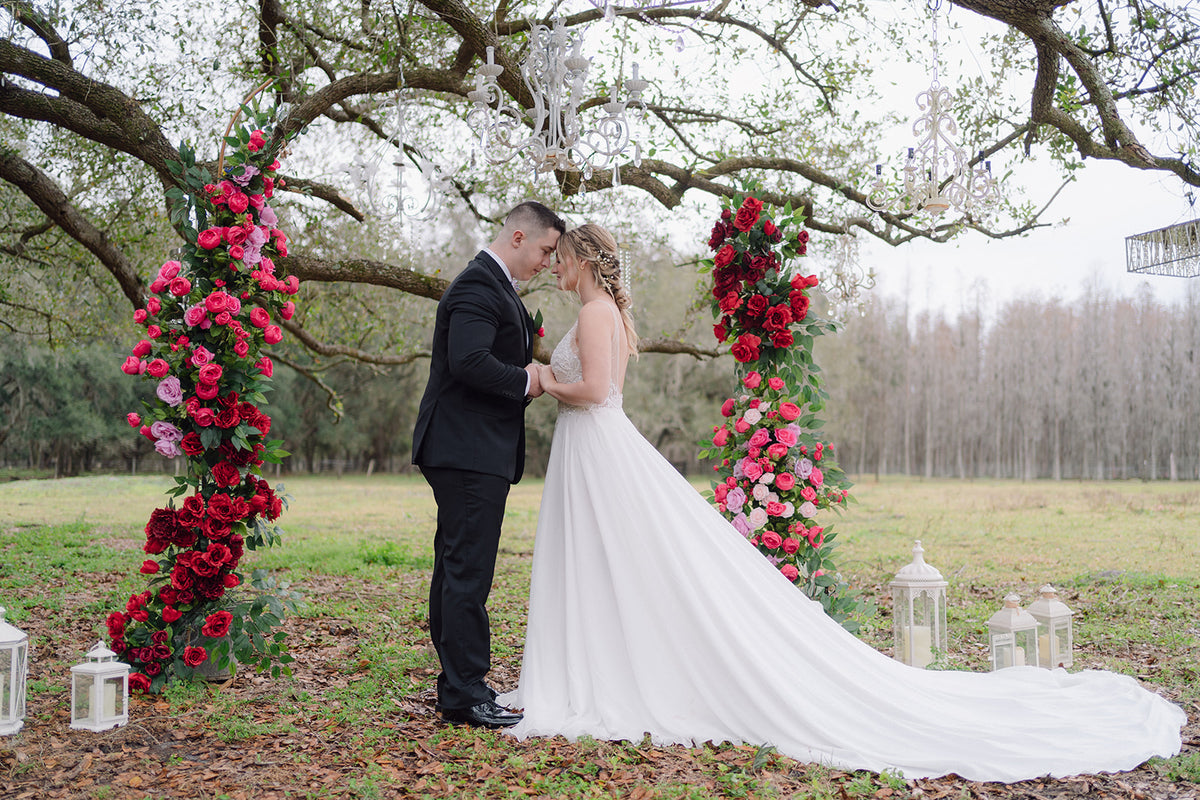 Couple sharing a kiss under a floral arch with red and pink flowers, surrounded by trees and lanterns.