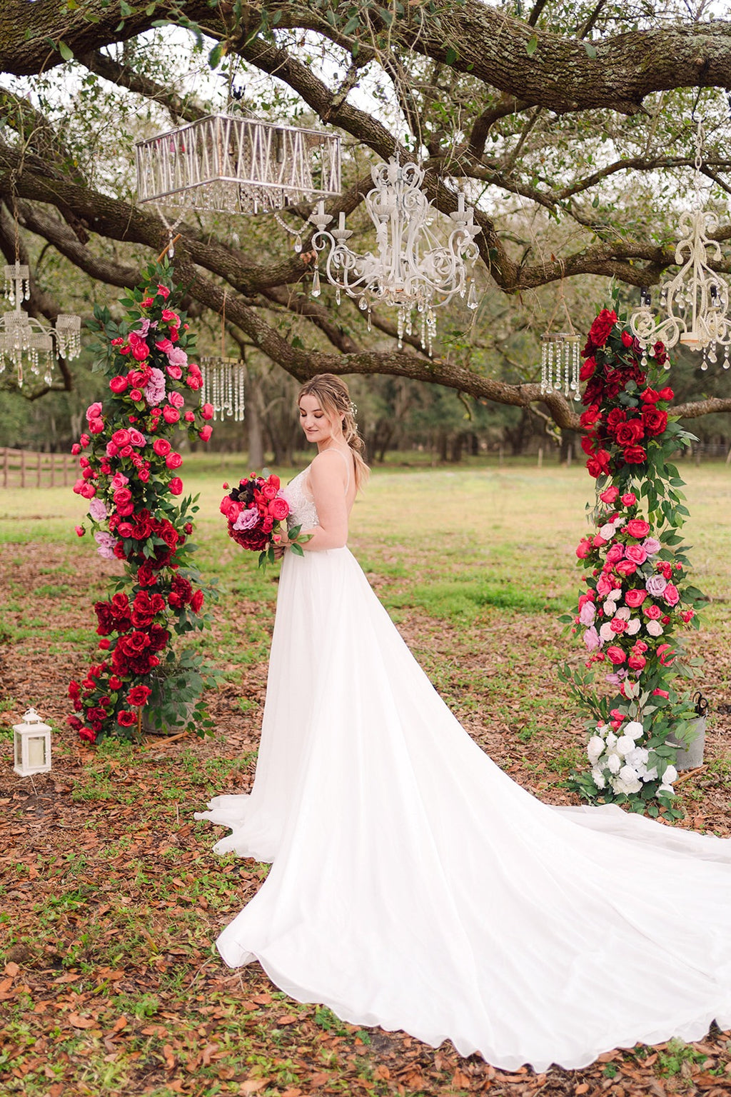 Bride standing under a decorated arch with red and pink artificial flower arrangements and chandeliers in an outdoor setting.