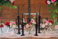Decorative table setting with black candles, small vases, and pink and red bud vase floral arrangements against a wooden background.
