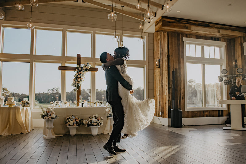 Groom lifting a bride in a wedding dress in a room with large windows and a cross with flower arrangement on the wall.