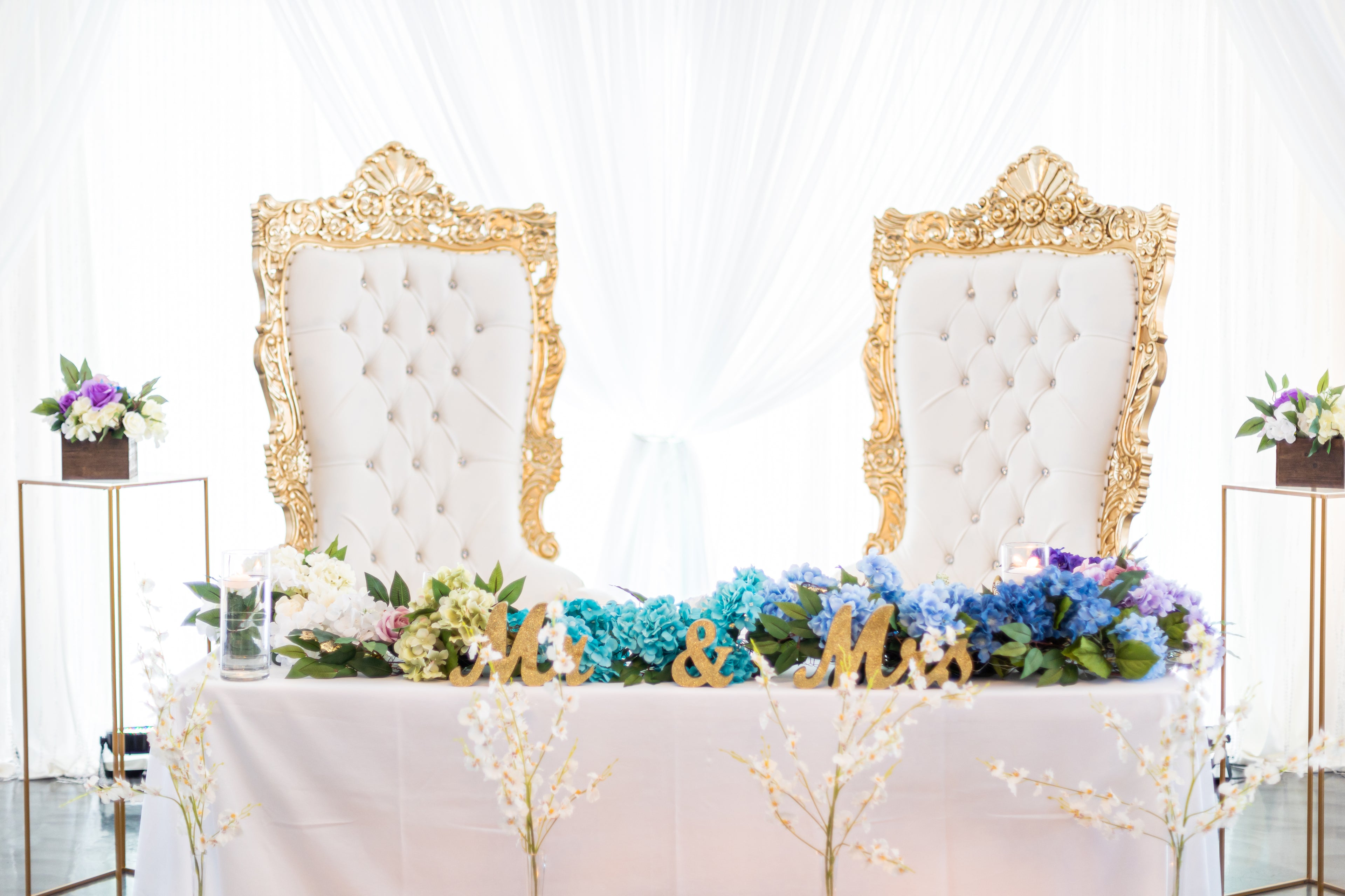 Decorated wedding sweetheart table with ornate chairs, purple and blue flowers, and 'Mr. & Mrs.' sign.