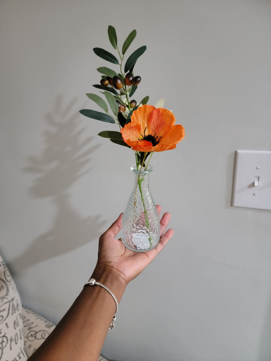 Hand holding a clear glass vase with an orange flower against a plain wall.